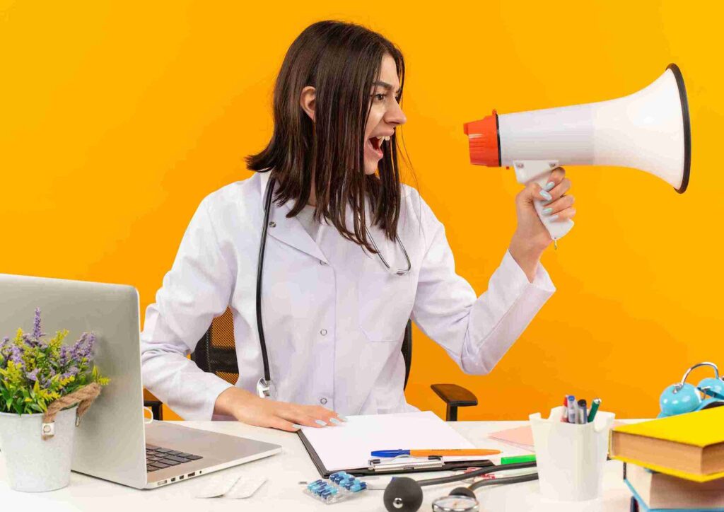 young-female-doctor-white-coat-with-stethoscope-shouting-megaphone-with-aggressive-expression-sitting-table-with-laptop-documents-orange-wal